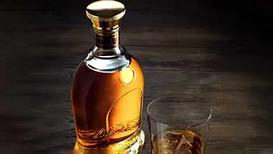 Ornate bottle of amber liquor with gold cap and base, next to a glass of the same liquor on the rocks, all on dark wood.