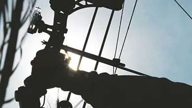 Silhouetted hand holding a compound bow, sun brightly behind it.