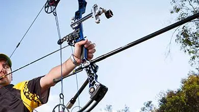 Low-angle, close-up of an archer aiming a blue and black compound bow against a clear sky.