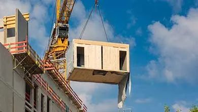A yellow crane lowers a wooden module onto a partially constructed building.