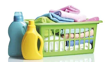 A green laundry basket filled with folded, colorful towels sits beside blue and yellow laundry detergent bottles on a white background.