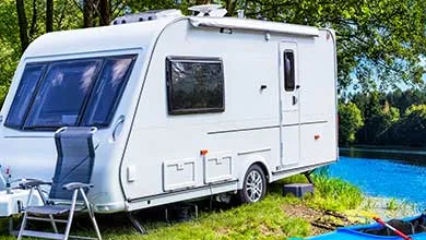 A white travel trailer is parked on grass by a blue lake, with a camping chair nearby and trees in the background.