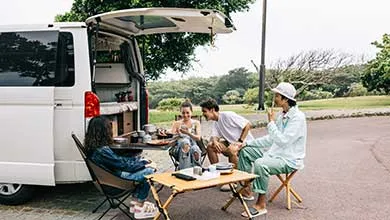 Four friends sit at folding tables beside a white camper van with its rear hatch open, eating and laughing together outdoors.