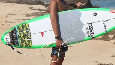 A surfer stands on a sandy beach holding a white surfboard with bright green edges, a gray hexagonal front pad, and a patterned tail pad.