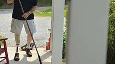 A man with a prosthetic leg sweeps a house’s front porch beside a red rocking chair.