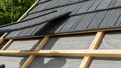 Close-up of dark gray roofing shingles being installed on a wooden roof frame, with one shingle lifted to show gray underlayment beneath; green trees in the background.