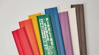 Eight colorful rectangular pipeline markers—red, orange, yellow, green, blue, purple, white, and dark brown—are fanned out against a plain light background. The green marker displays bold vertical text reading "CAUTION PIPELINE," with "811" and additional symbols below.